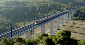 La imagen muestra un tren de alta velocidad circulando sobre un viaducto elevado en una zona montañosa. El tren es de color rojo con detalles en gris y está compuesto por varios vagones aerodinámicos. El viaducto está sostenido por pilares de hormigón altos y atraviesa un paisaje natural con abundante vegetación, principalmente pinos, en el fondo y en las laderas. El cielo no es visible, pero la iluminación indica que es de día, con luz natural clara.