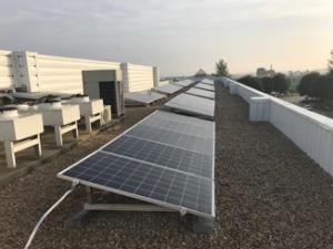 Paneles solares instalados en el tejado de un edificio industrial, colocados en fila sobre una superficie de grava. Al fondo se observan equipos de climatización y un paisaje con árboles y cielo despejado.