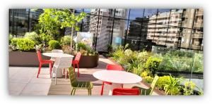 Terraza con mesas y sillas de colores rodeada de plantas, con edificios reflejados en una pared de vidrio.