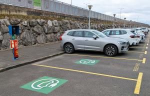 Aparcamiento con varias plazas para coches eléctricos, con señalizaciones verdes y un punto de carga junto a los vehículos estacionados.