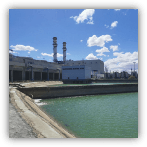 Vista de una planta industrial junto a un canal de agua verde, con dos chimeneas altas al fondo bajo un cielo azul con nubes.