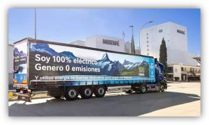 Camión eléctrico con mensaje sobre cero emisiones estacionado frente a una fábrica industrial.