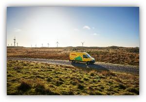 Furgoneta de reparto amarilla circulando por un camino rural con aerogeneradores al fondo.