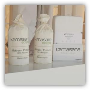 Tres botes blancos y dos cajas de protectores de colchón Kamasana sobre una mesa blanca.