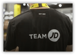 Parte trasera de una camiseta negra con el logotipo "Team JD" en un entorno comercial o logístico.