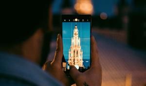 En el centro de la imagen aparece un teléfono móvil sostenido con ambas manos. La cámara del móvil está abierta y enfocando una torre o campanario iluminado. La fotografía está tomada desde detrás de la persona que sostiene el dispositivo, creando una composición de "foto dentro de la foto".