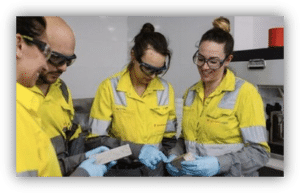 Cuatro personas con ropa de protección de alta visibilidad, gafas de seguridad y guantes examinan varias muestras en un laboratorio.