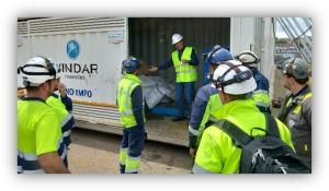 Grupo de trabajadores con chalecos reflectantes y cascos de seguridad participando en una sesión informativa junto a un contenedor de almacenamiento de hidrógeno en una instalación industrial.
