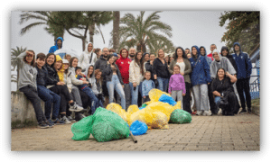 Grupo de personas posando junto a varias bolsas de residuos recogidos durante una actividad de limpieza en una zona ajardinada con palmeras.
